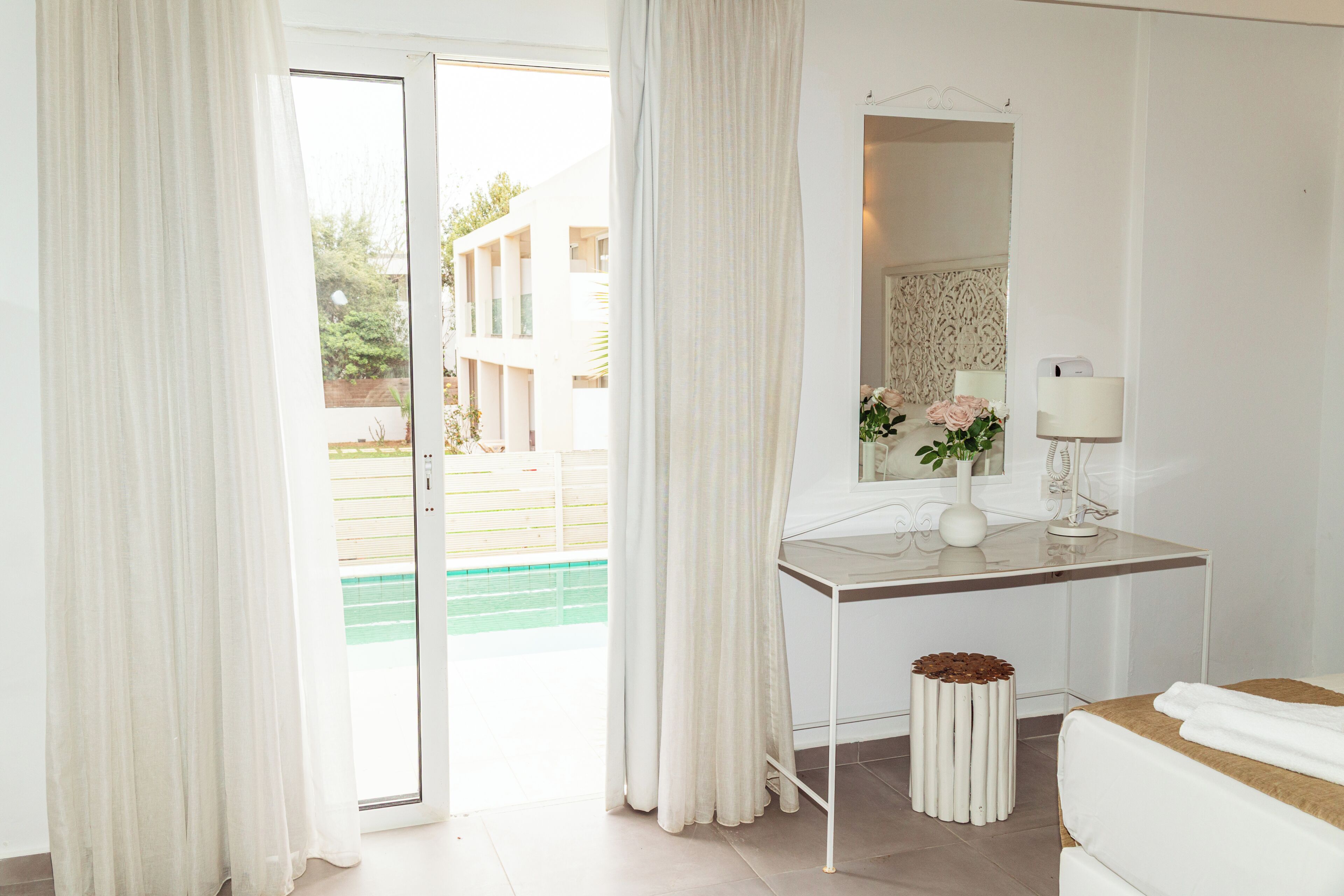 Habitación de hotel con cama, escritorio con espejo, lámpara y vistas a una piscina y edificios.