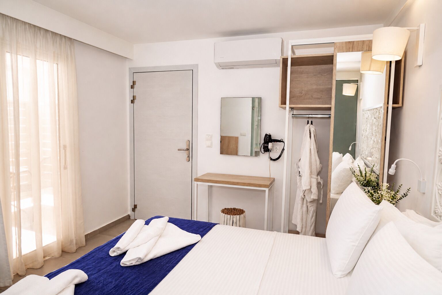 A hotel room with a bed, a vanity with a mirror, a towel rack, and a window with curtains.