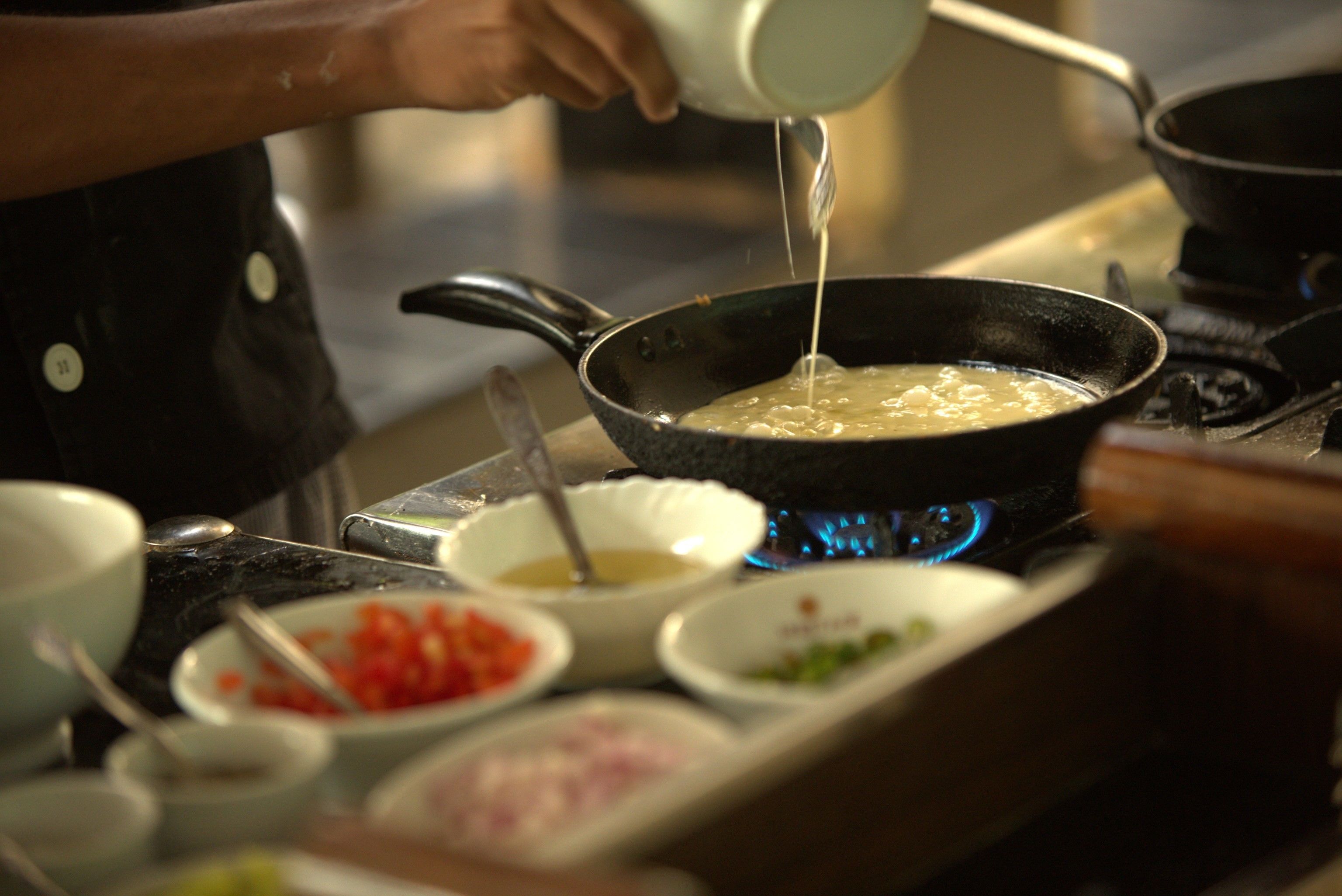 A person cooking in a kitchen with a pan on a stove, various bowls with ingredients, and a pot on a blue flame.