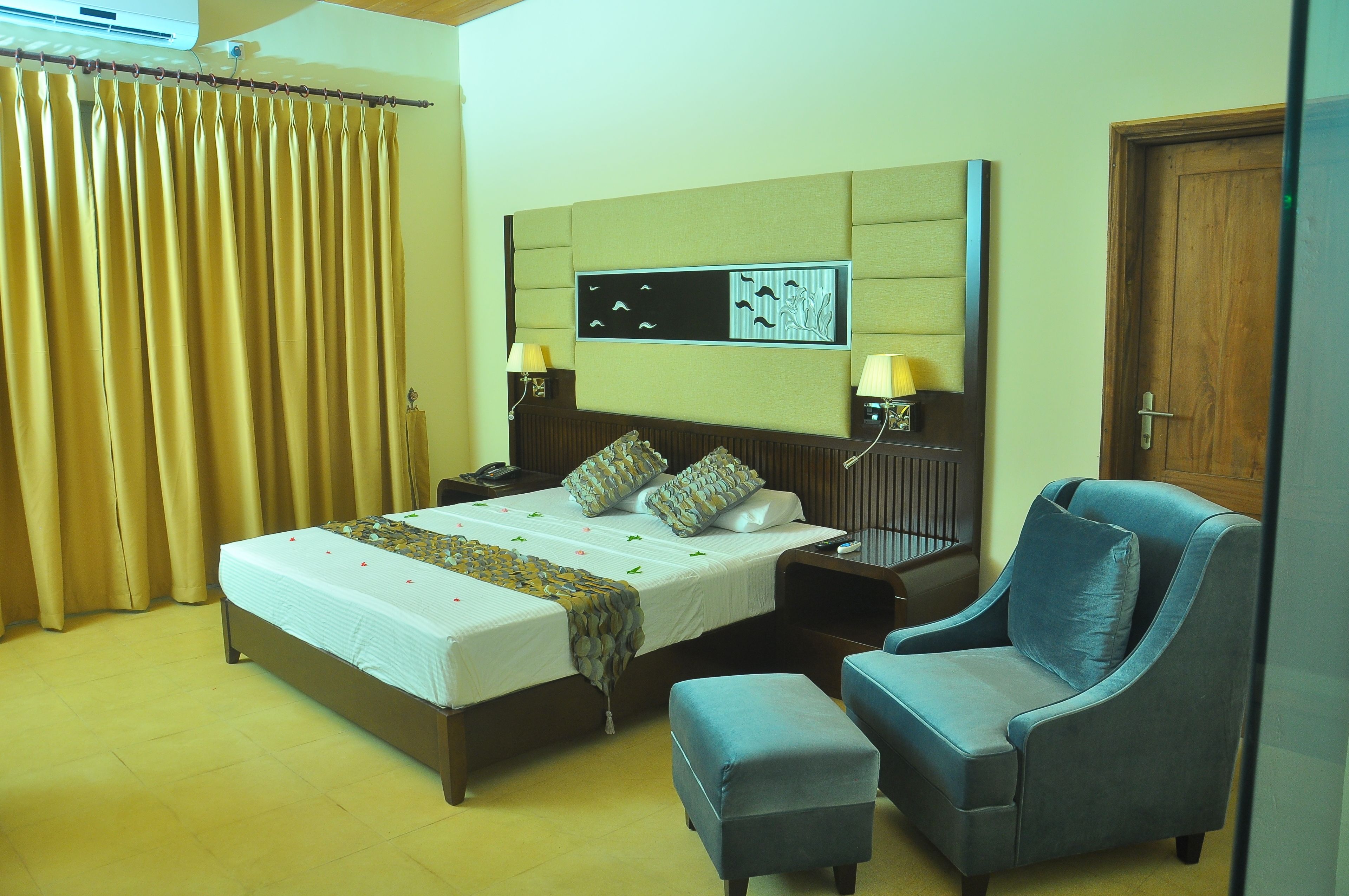 A hotel room with a bed, two bedside tables with lamps, a chair, an ottoman, and a wall-mounted artwork.