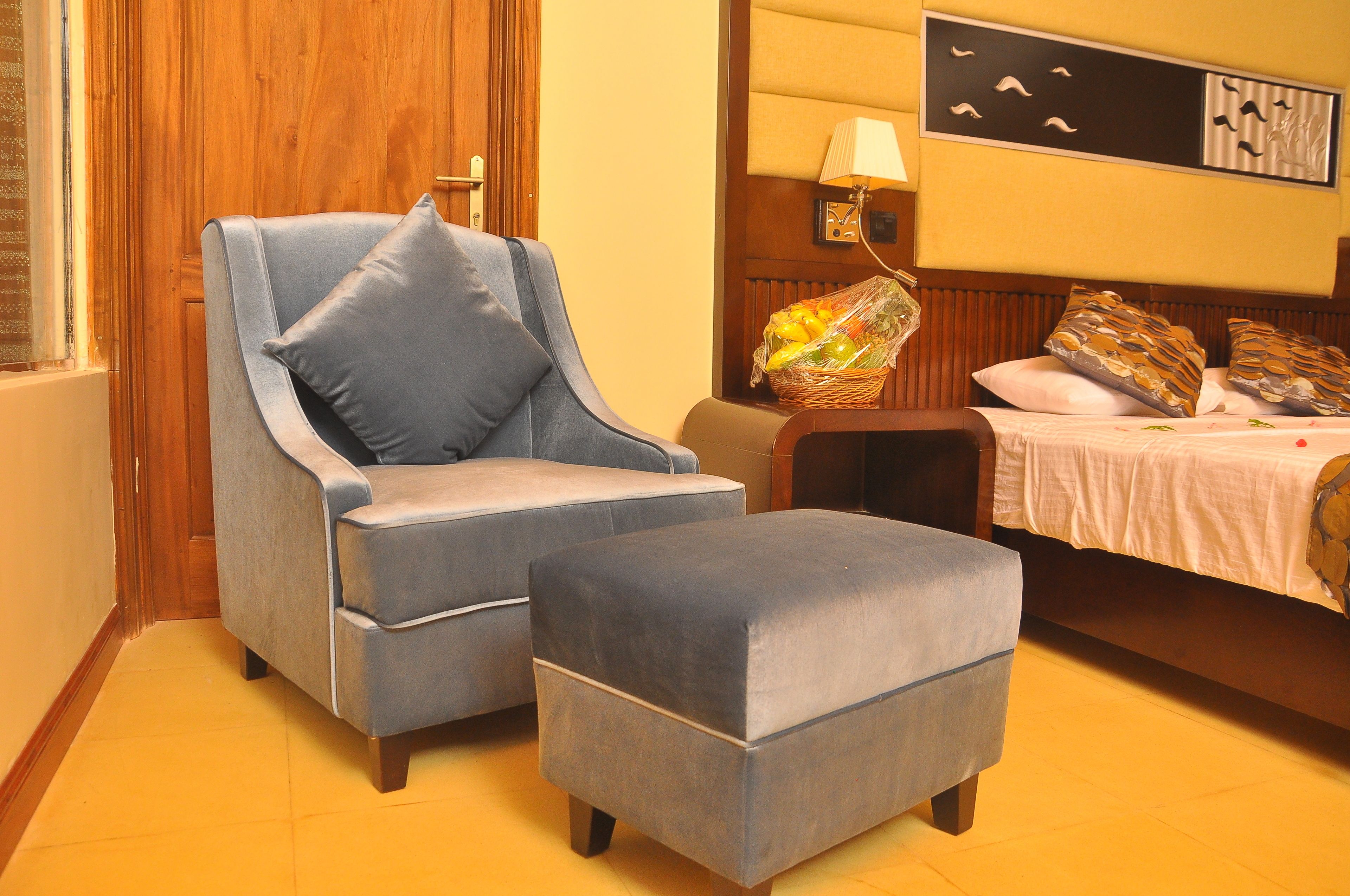 Una habitación con puerta de madera, un sillón azul con su reposapiés a juego, una mesita con una cesta de fruta y una cama con almohadones estampados.