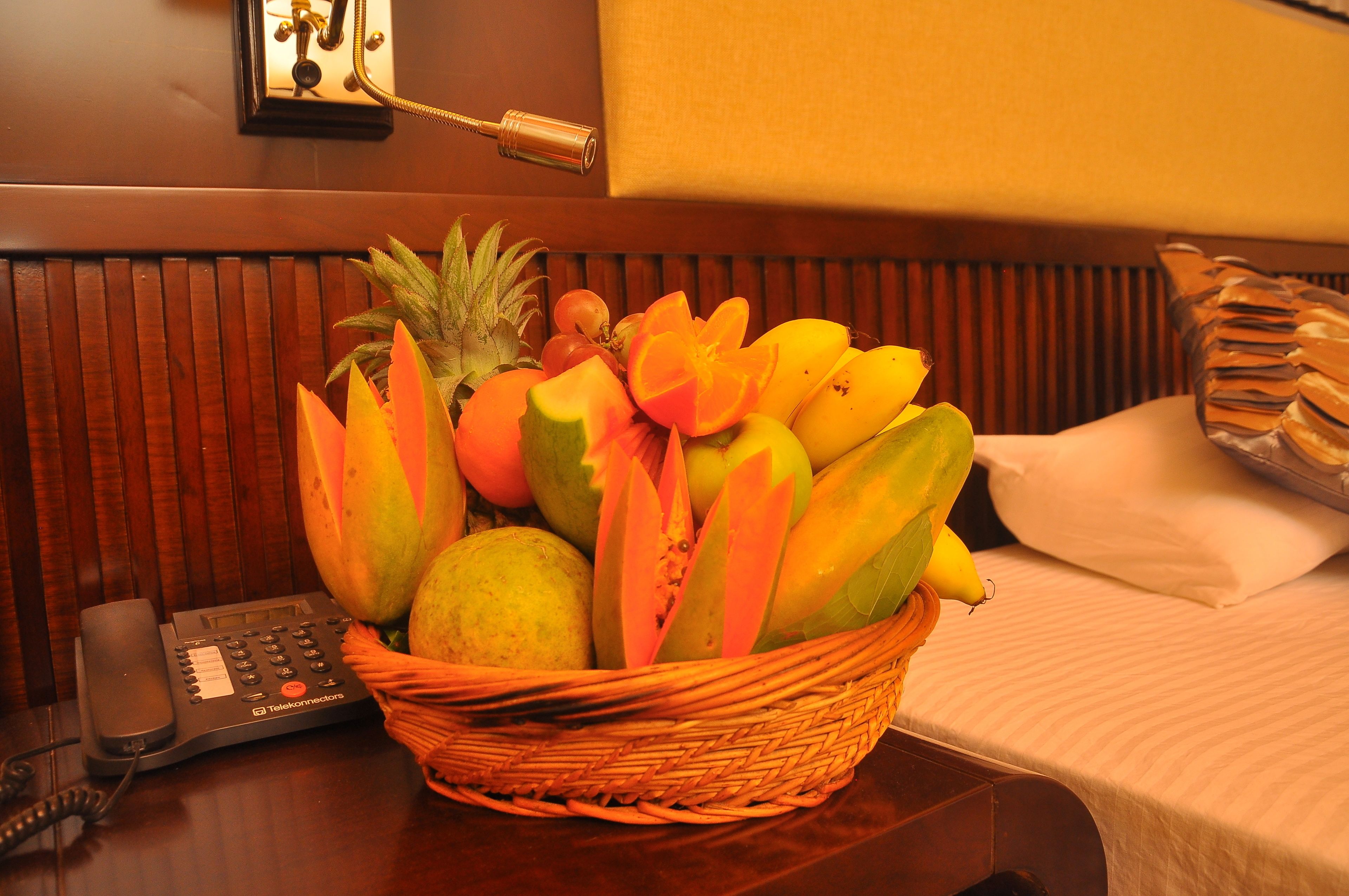 A fruit basket on a hotel room table with a telephone and a wall clock.