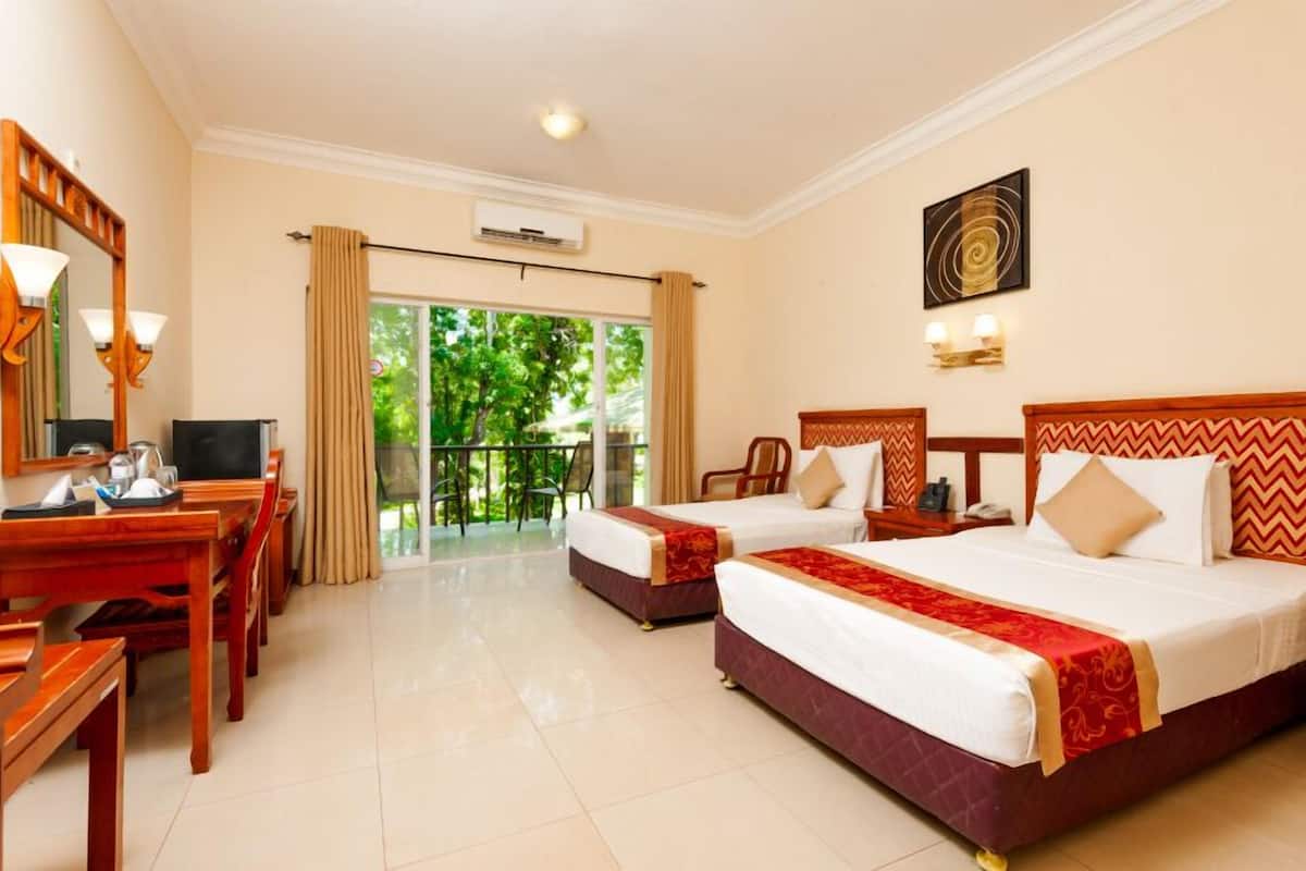 A hotel room with two beds, a desk, a chair, and a balcony with a view of greenery.