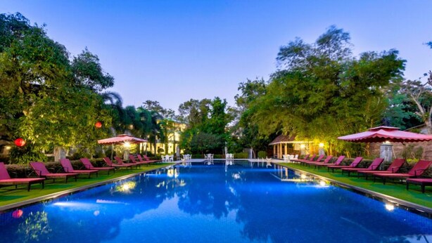 A swimming pool with lounge chairs and umbrellas, surrounded by trees and a building with lights. - Heritage Hotel (Anuradhapura)