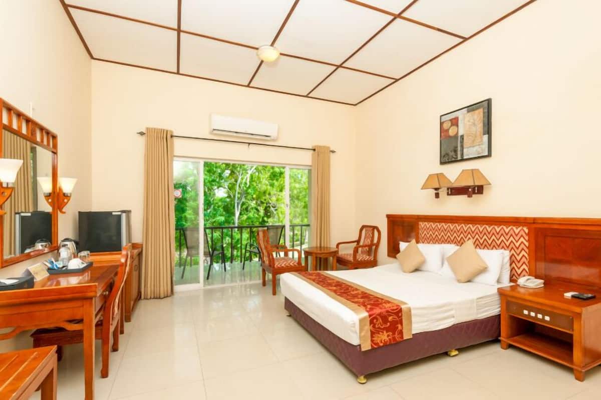 A hotel room with a bed, a desk with a TV, a balcony with a view of greenery, and a painting on the wall.
