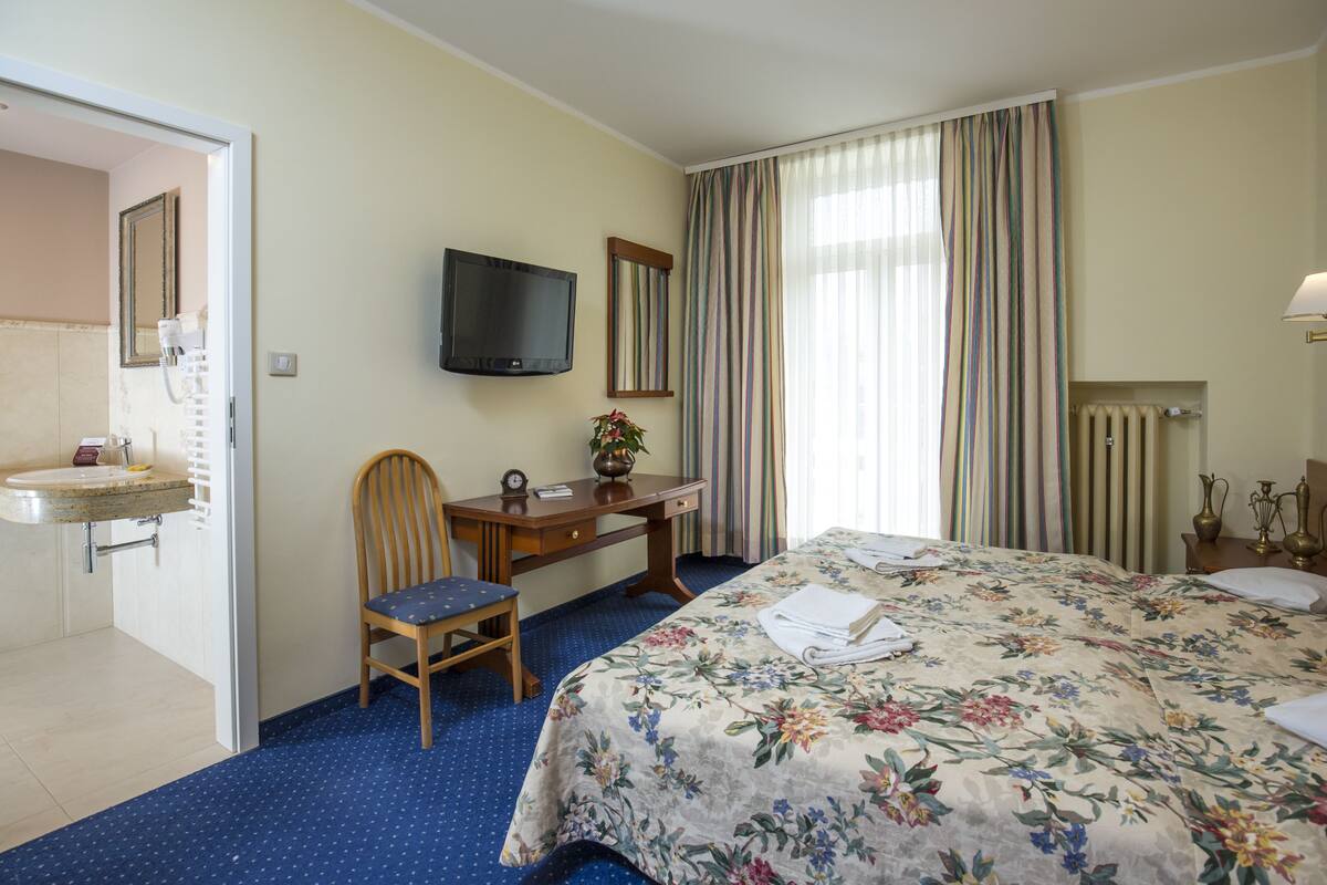 Habitación de hotel con cama, escritorio, silla, televisión, baño con lavamanos y ventana con cortinas.