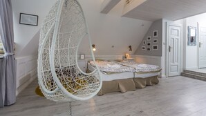 Une chambre avec un fauteuil suspendu, un lit recouvert d’une couvre-lit beige, une table de chevet avec une lampe et un mur orné de tableaux encadrés.