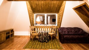 Une pièce mansardée chaleureuse avec un plafond en bois, une petite table recouverte d’une nappe à motifs, deux chaises en bois, un canapé violet et un banc en bois.