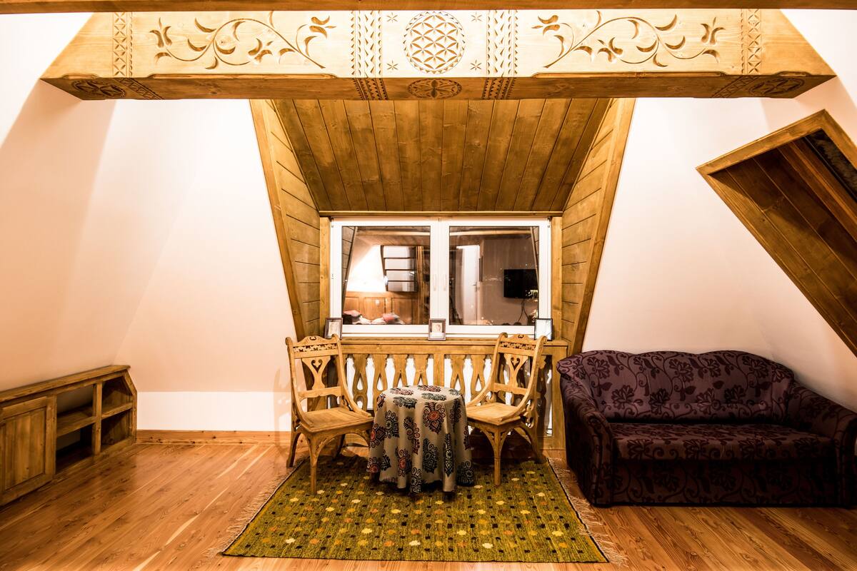 Una habitación cálida en el ático con techo de madera, una pequeña mesa con mantel estampado, dos sillas de madera, un sofá morado y un banco de madera.