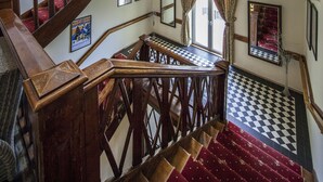 Un escalier en bois avec un sol à carreaux, menant à un palier recouvert d’un tapis rouge.