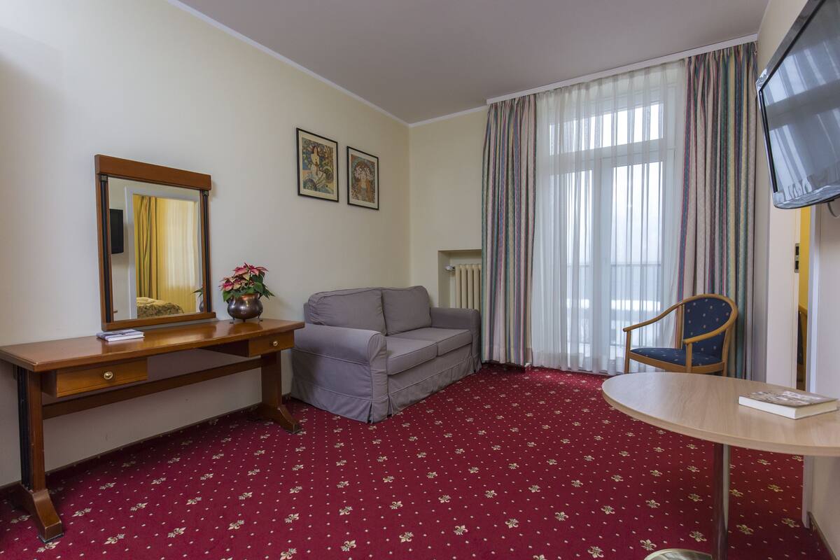 Habitación de hotel con un sofá gris, un escritorio de madera con un espejo, una mesita pequeña, una silla azul y un televisor.
