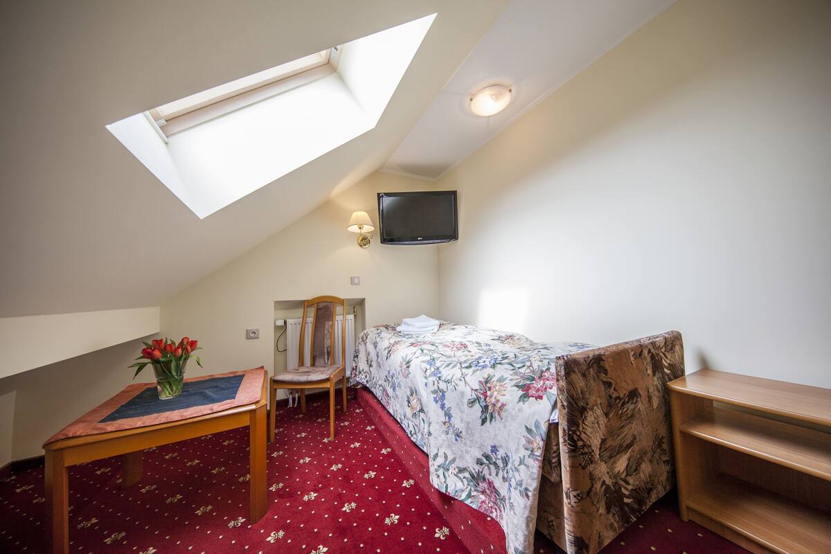 Una habitación pequeña y acogedora con techo inclinado, una cama individual, una silla de madera, una mesita con un jarrón de flores y un televisor montado.