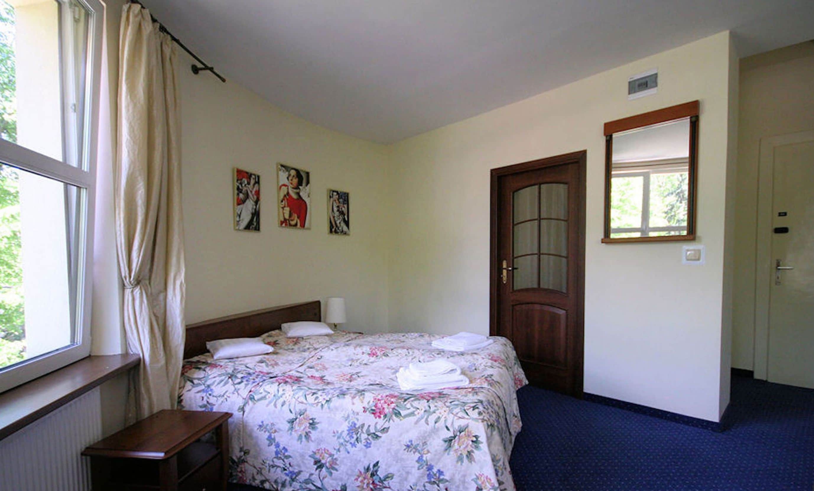 Un dormitorio con cama, mesita de noche, ventana con cortinas, puerta y tres cuadros en la pared.