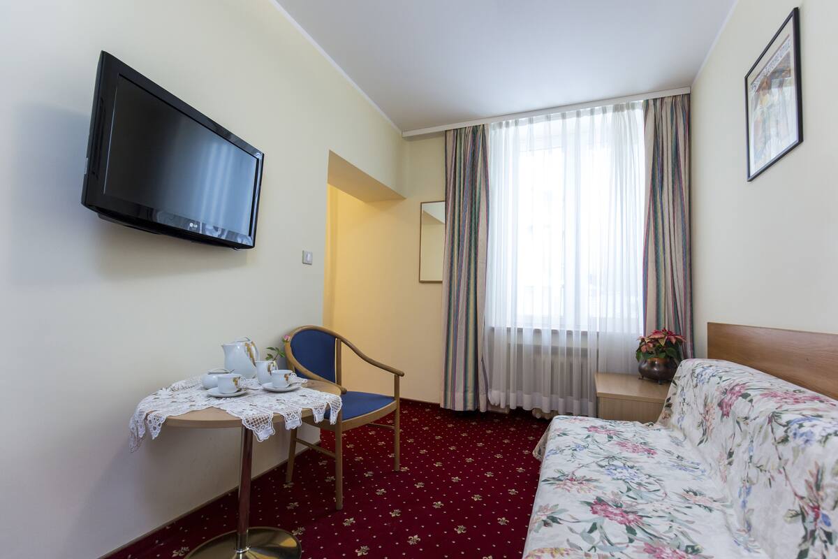 Habitación de hotel con una cama, un escritorio con silla, un televisor en la pared y una ventana con cortinas.