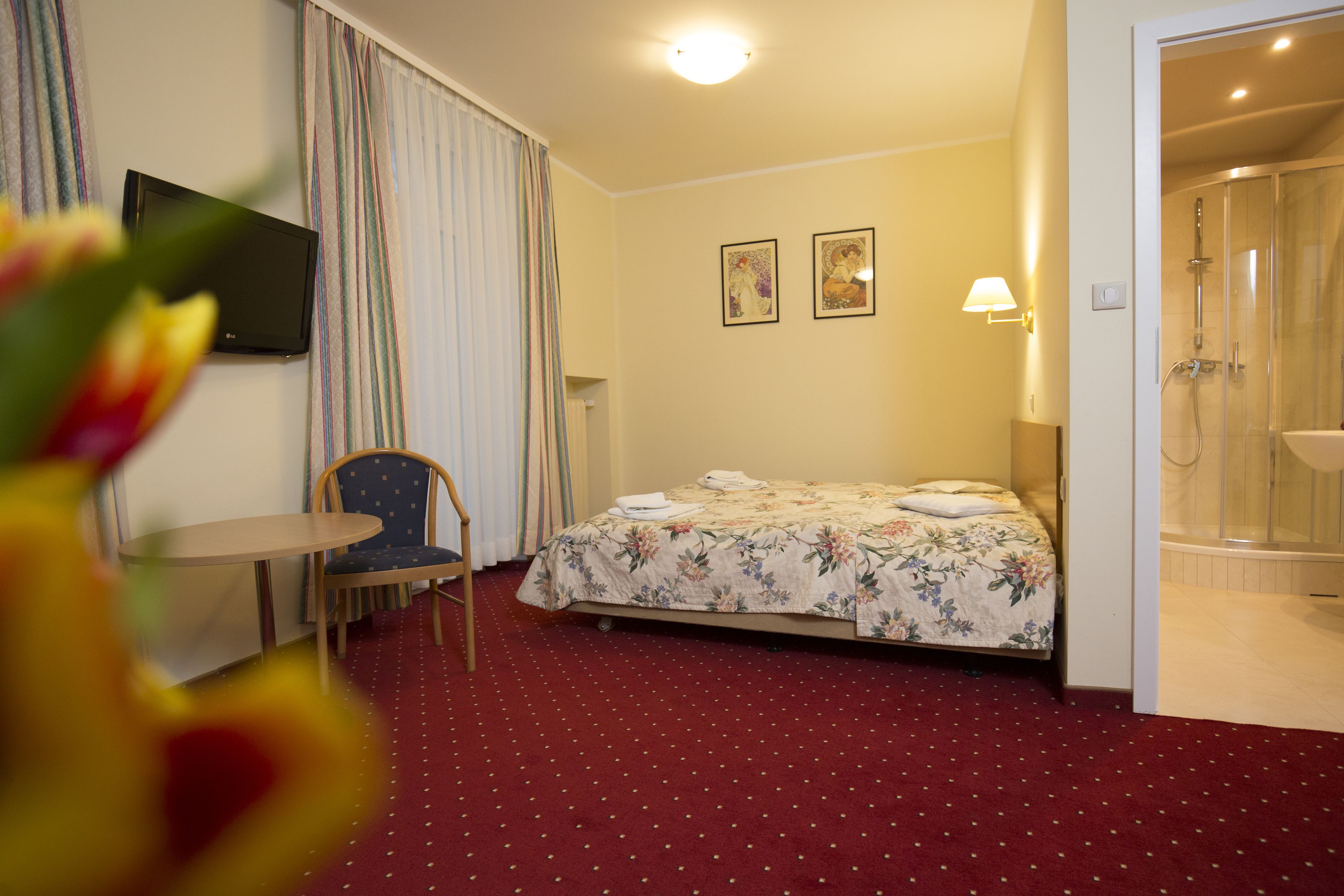 A hotel room with a bed, a chair, a small table, a shower area, and a TV on the wall.
