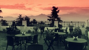 Un café al aire libre con mesas y sillas redondas, rodeado de plantas en macetas y con vistas a un paisaje montañoso.