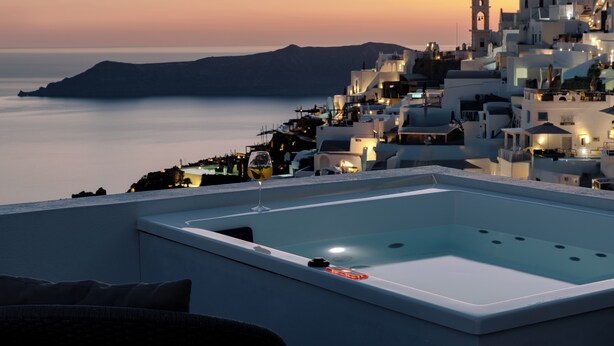 Une piscine sur le toit, un verre de vin à la main, avec vue sur une ville côtière aux bâtiments blancs et un coucher de soleil.