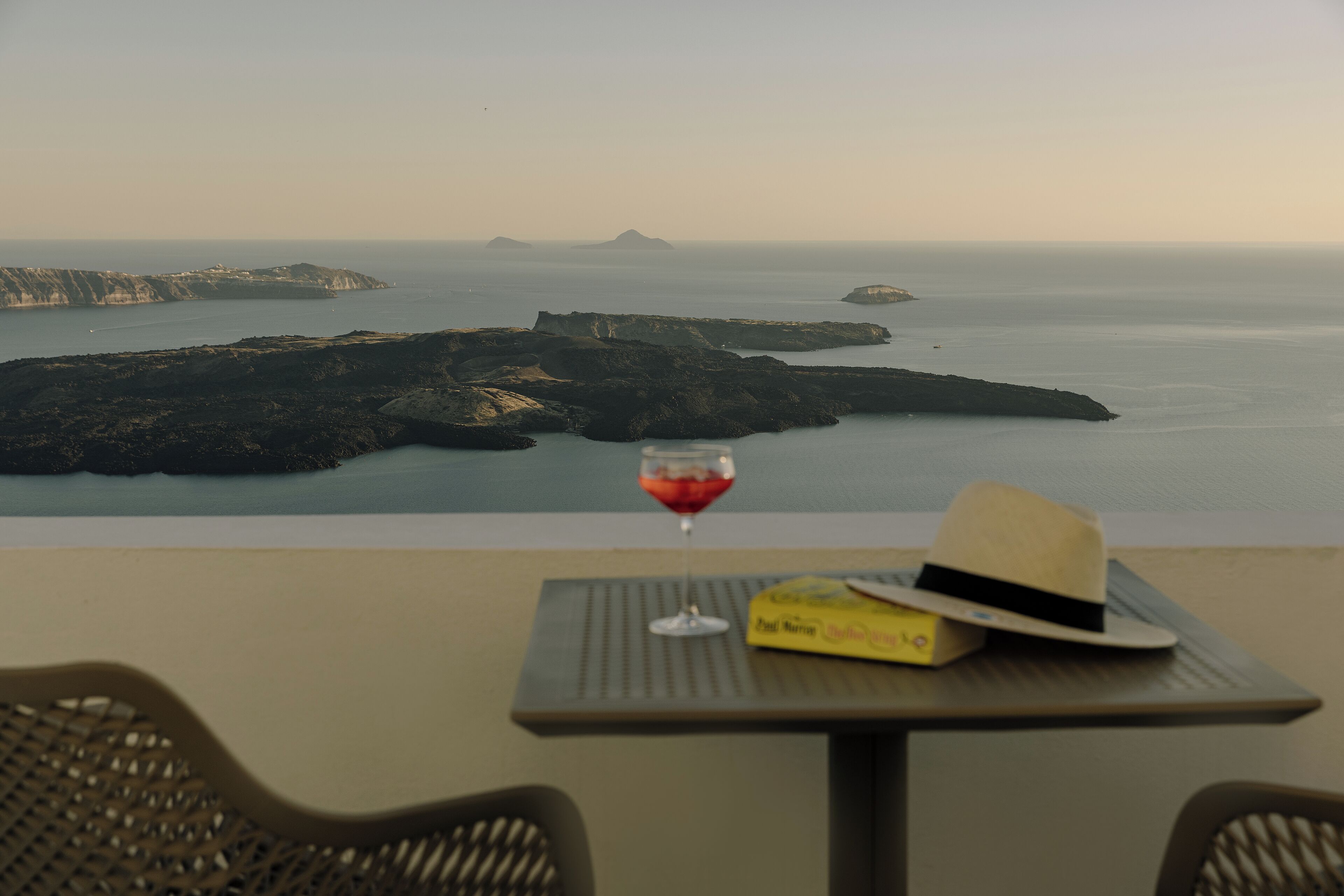 A table with a hat and a glass of red wine, overlooking a coastal landscape with cliffs and calm sea.