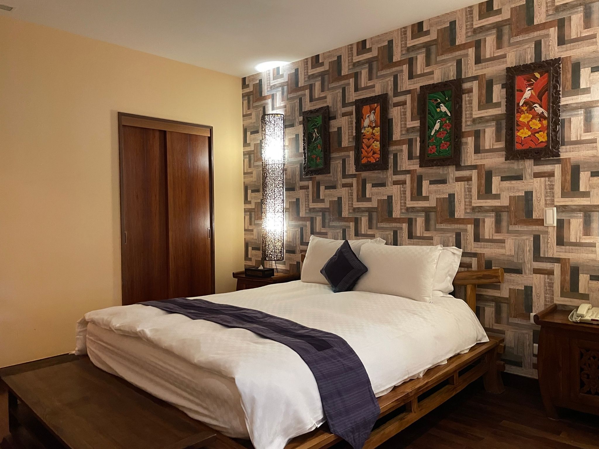 A bedroom with a wooden bed, a nightstand, a lamp, and a wall with a geometric pattern.