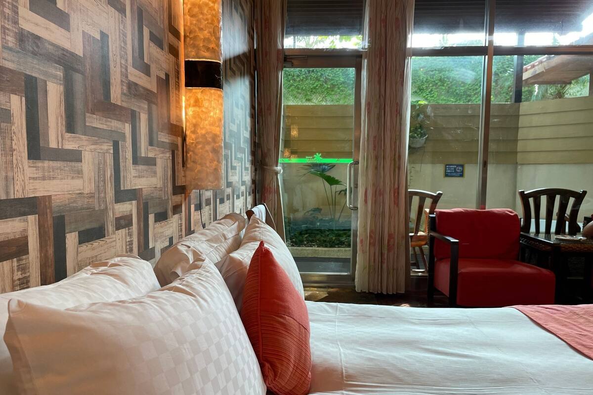 A hotel room with a bed, a red chair, and a view of greenery outside.