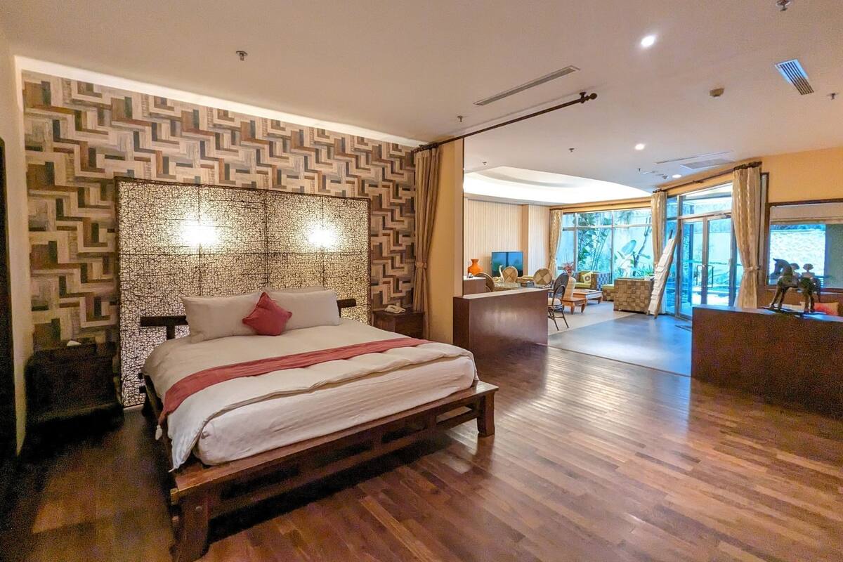 A modern hotel room with a large bed, a wooden headboard, and a view of the outdoors through large windows.
