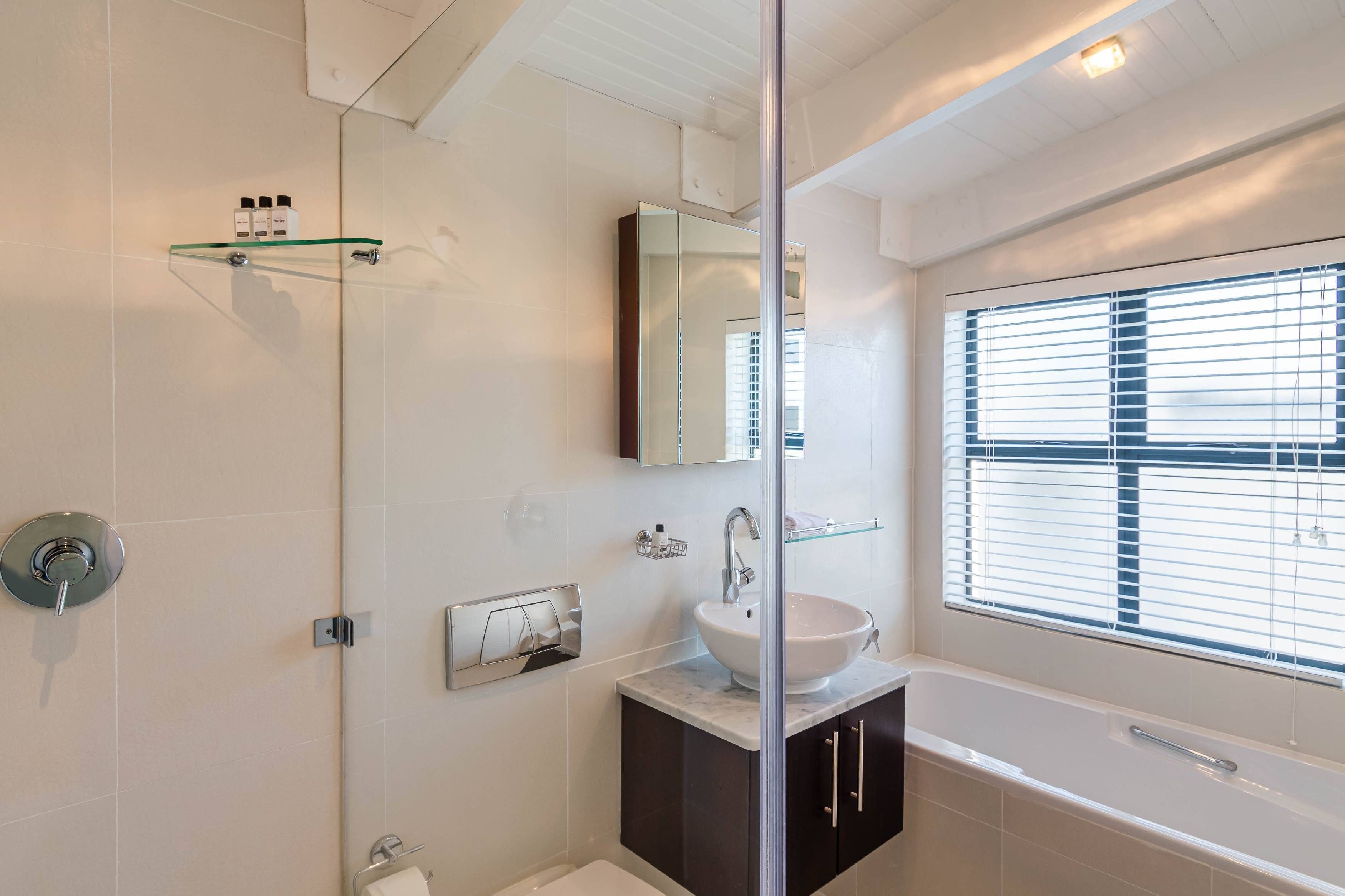 A modern bathroom with a glass shower enclosure, a white sink, dark cabinets, and a large window with blinds.