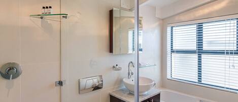 A modern bathroom with a glass shower enclosure, a white sink, dark cabinets, and a large window with blinds.