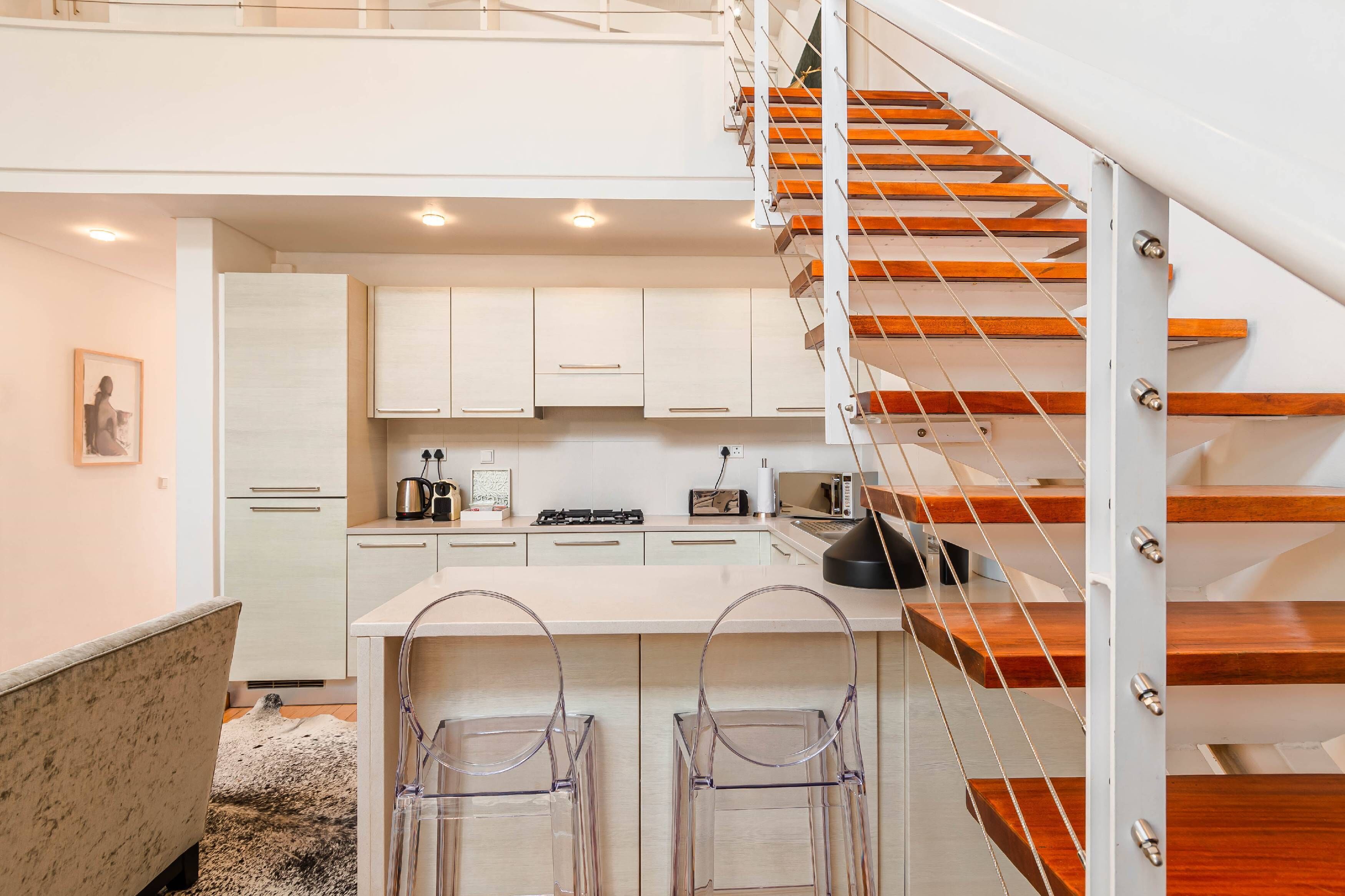 A modern kitchen with white cabinets, a wooden staircase, and a bar area with transparent chairs.