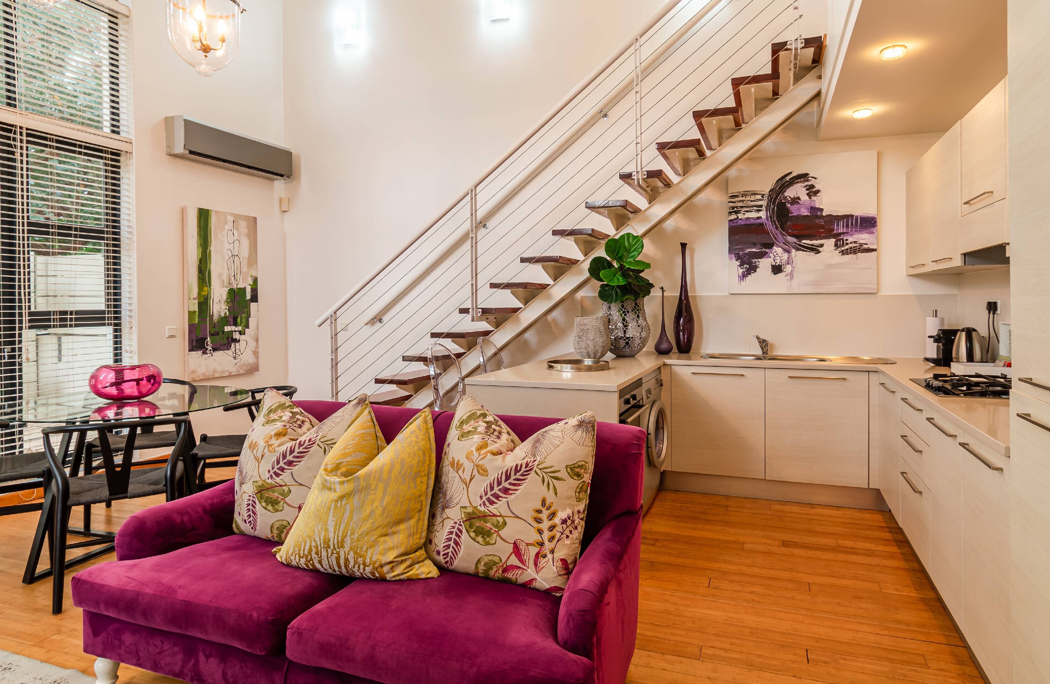 A modern living room with a purple sofa, a staircase, and a kitchen area.