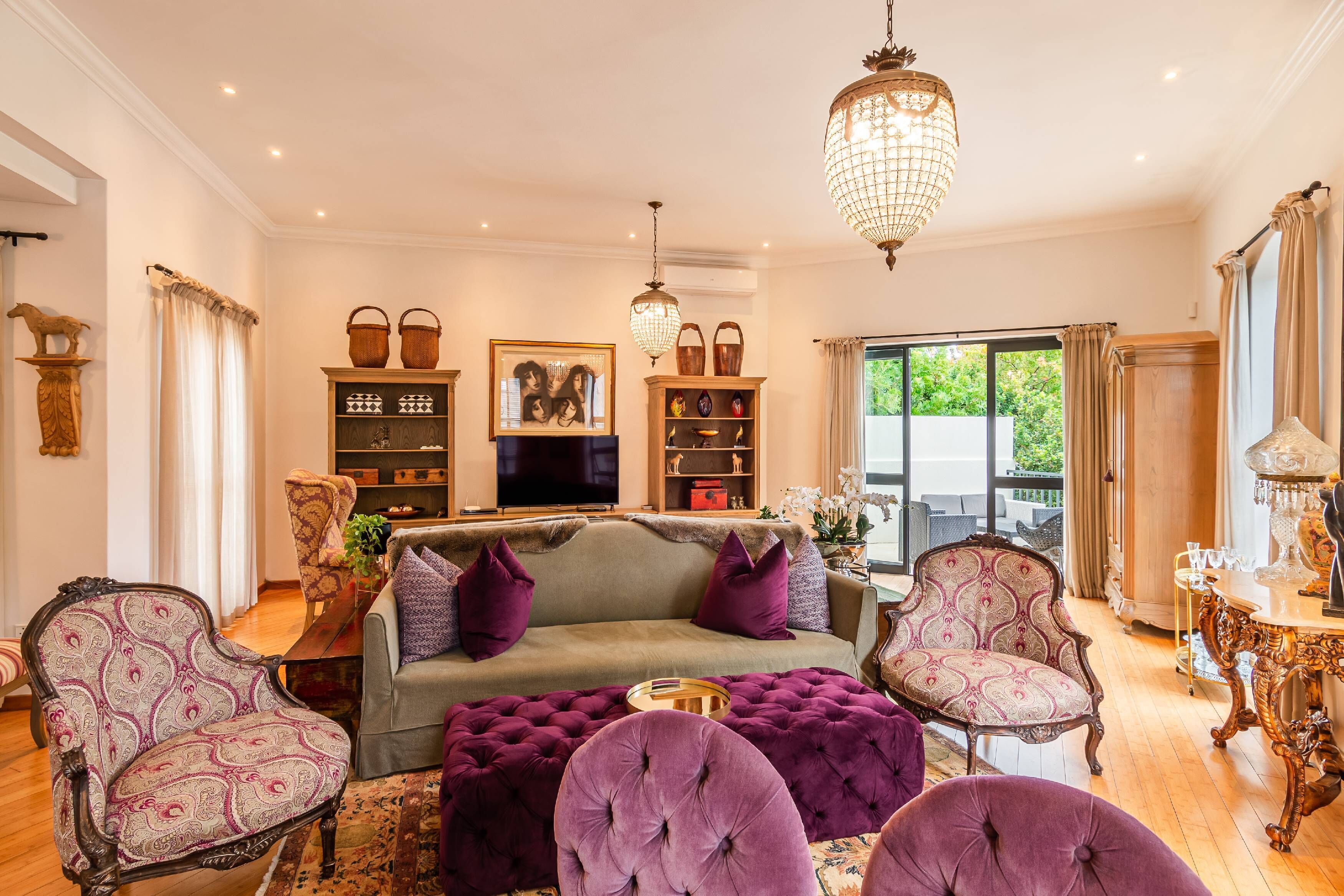 A living room with a sofa, armchairs, a coffee table, and a chandelier. There are shelves with decorative items and a television.