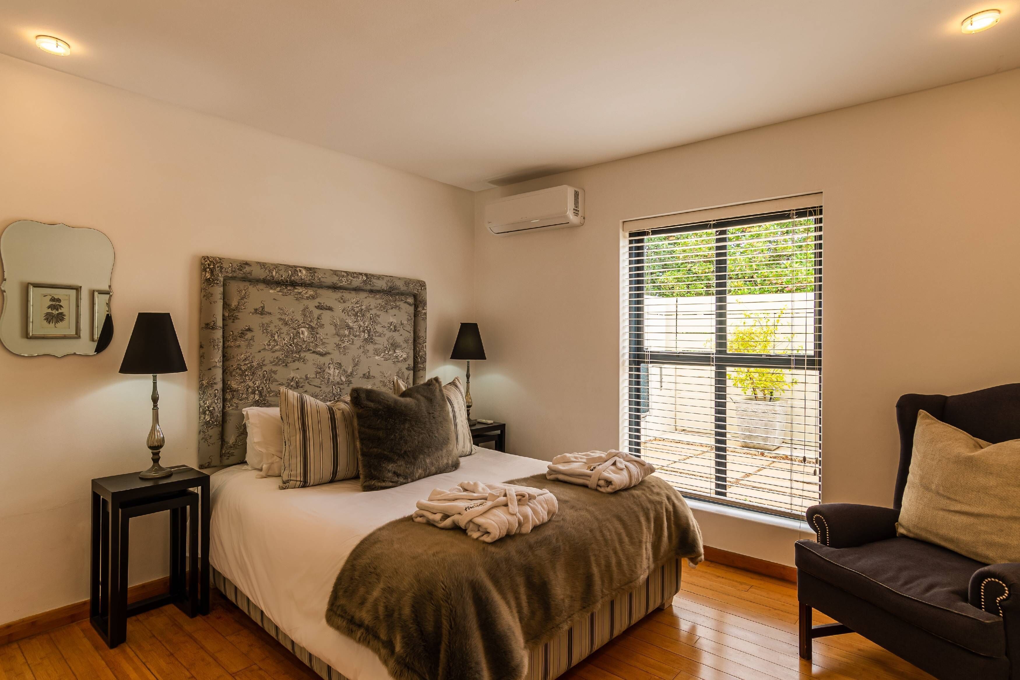 A bedroom with a large bed, two bedside tables, a nightstand, a window with blinds, and a chair.