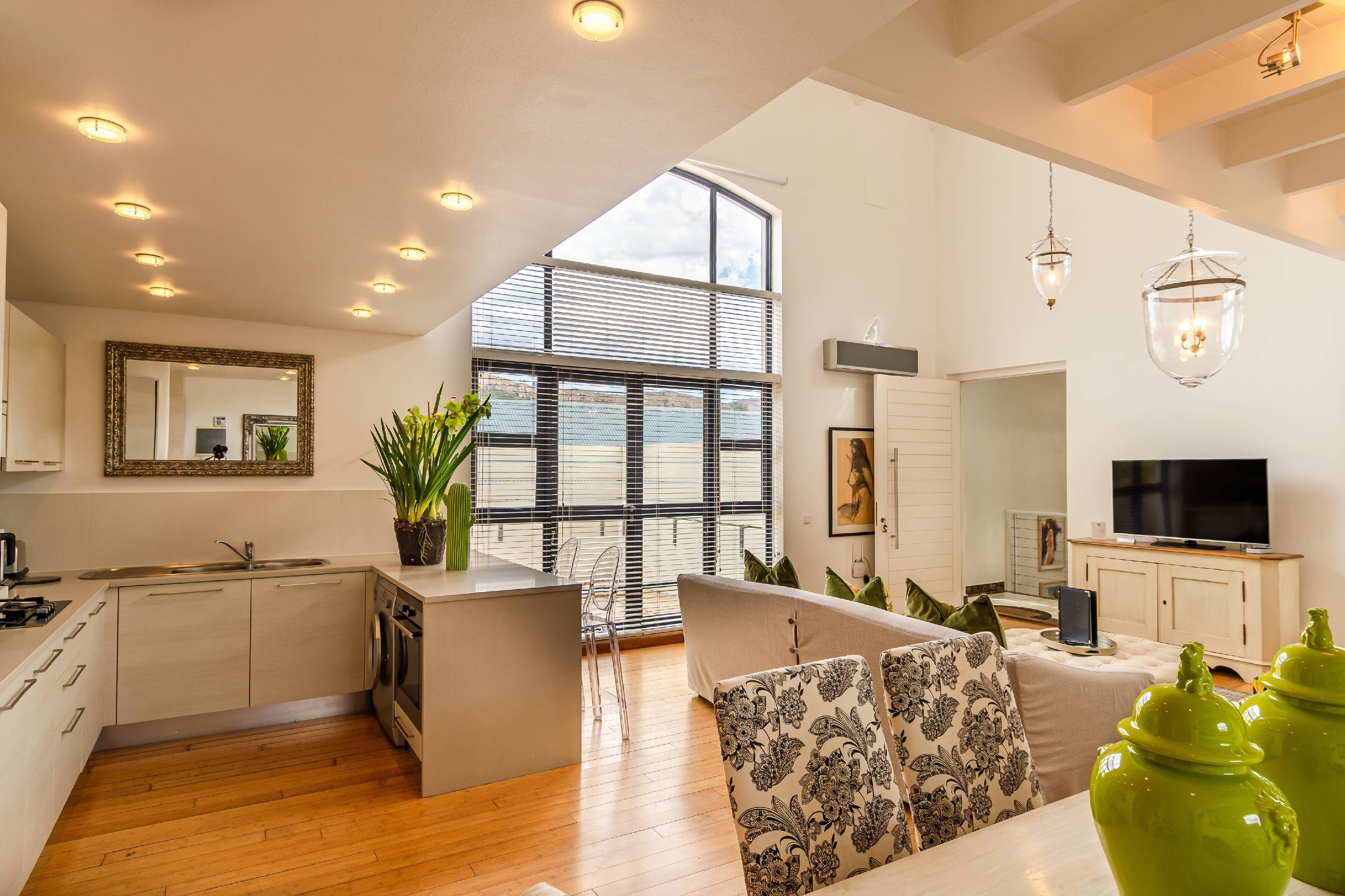 A modern kitchen with a central island, a dining area with patterned cushions, and a large window with blinds.