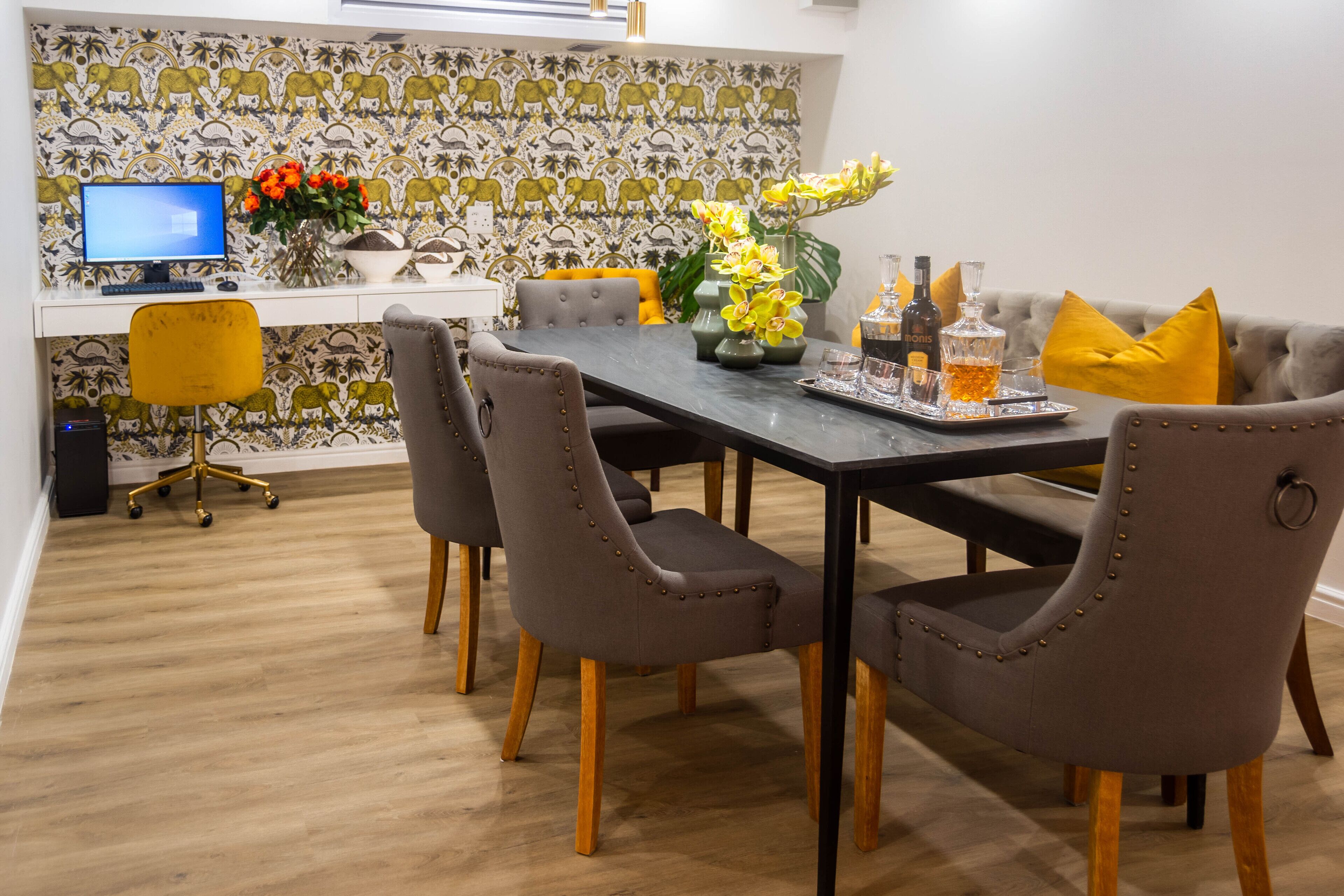 A dining area with a long table, four chairs, a computer on a desk, and a decorative wall with a pattern.