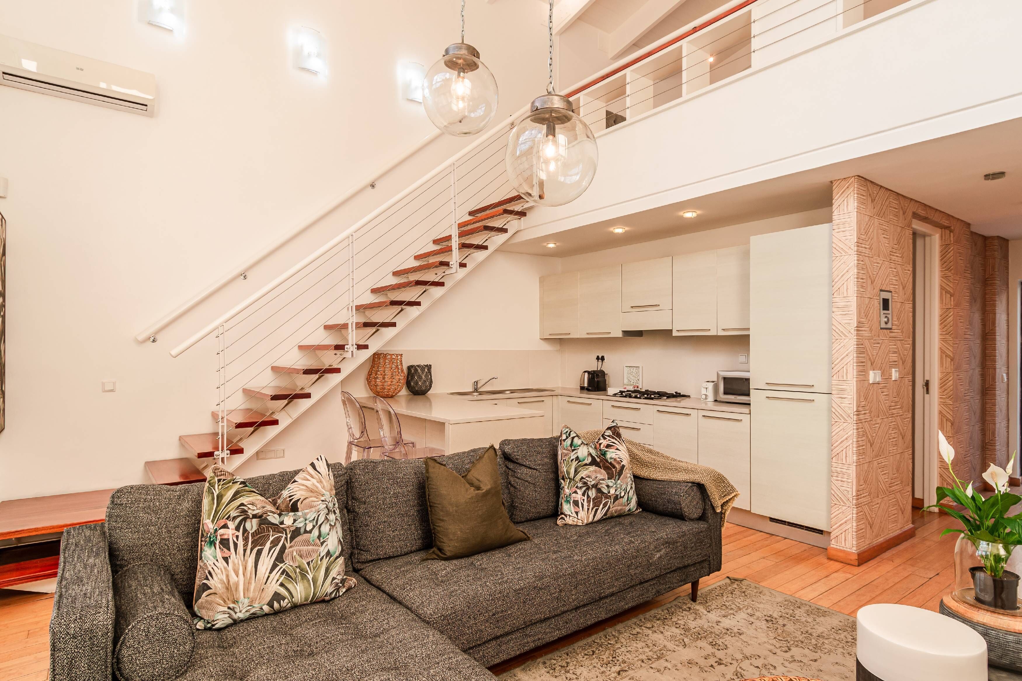 A modern living room with a grey sectional sofa, a staircase, and a kitchen area.