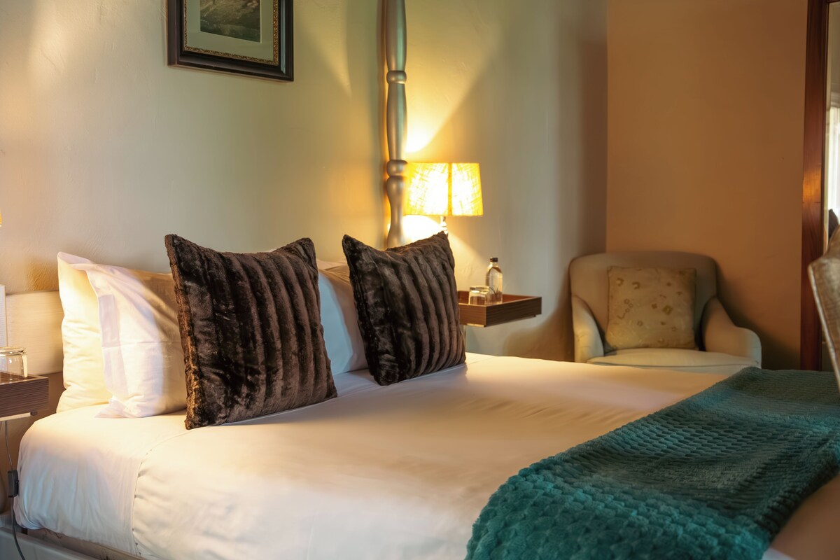 A hotel room with a bed, pillows, a chair, a nightstand, a lamp, and a picture on the wall.
