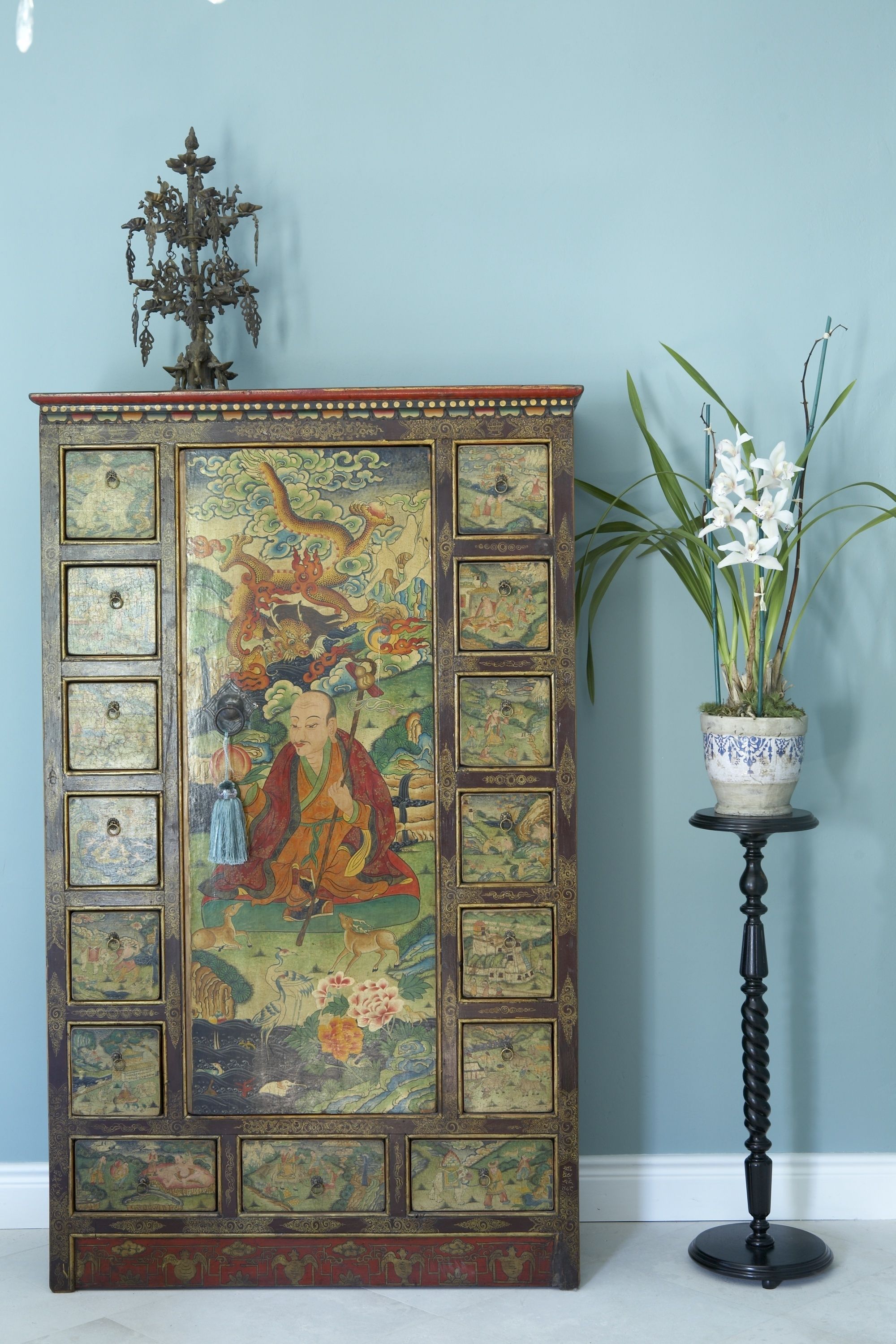 Un mueble tradicional con intrincados detalles decorativos, una mesita con una planta en maceta y una lámpara ornamental.