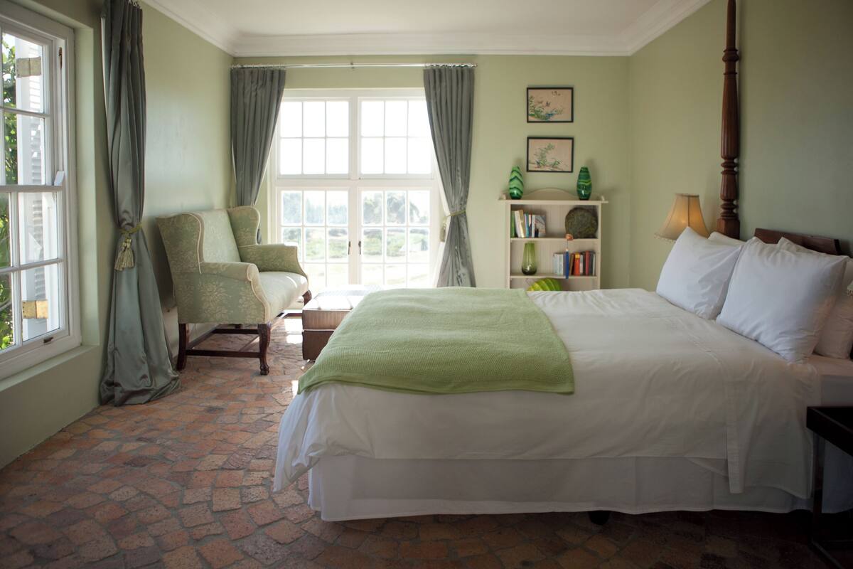 Ein Schlafzimmer mit einem großen Bett, einem Sessel, einem Bücherregal und einem Fenster mit Vorhängen.