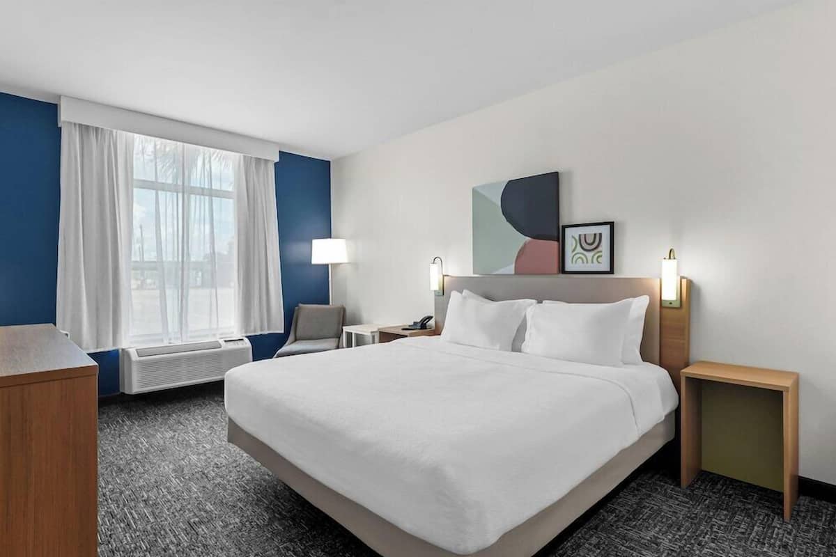 A hotel room with a large bed, bedside tables, a desk, a chair, a window with curtains, and a wall-mounted artwork.