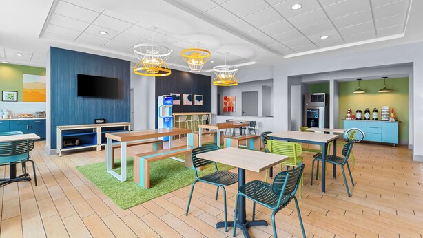 A modern, well-lit dining area with wooden tables and green chairs, a flat-screen TV, and a kitchen area with a coffee machine. - Spark by Hilton Victoria (Victoria)