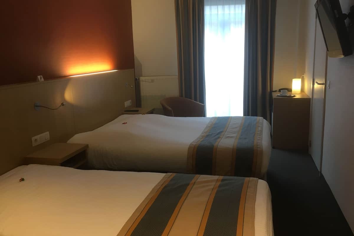Ein Hotelzimmer mit zwei Betten, einem Schreibtisch, einem Stuhl, einem Fernseher und einem Fenster mit Vorhängen.