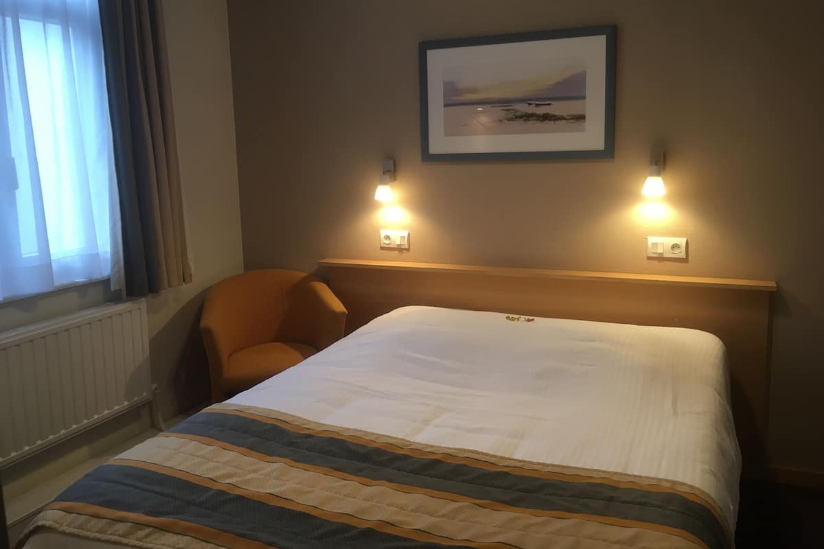 Ein Hotelzimmer mit einem Bett, einem Sessel, einem Fenster mit Vorhängen und zwei Wandleuchten.
