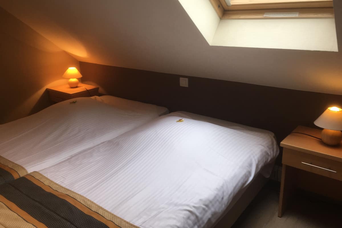 Ein Einzelbett mit weißer Matratze und einem kleinen Nachttisch mit Lampe. Über dem Bett ist ein Dachfenster zu sehen.