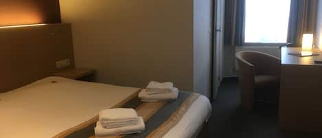 Ein Hotelzimmer mit einem ordentlich bezogenen Bett, einem Schreibtisch mit Lampe und einem Fenster mit Vorhängen.