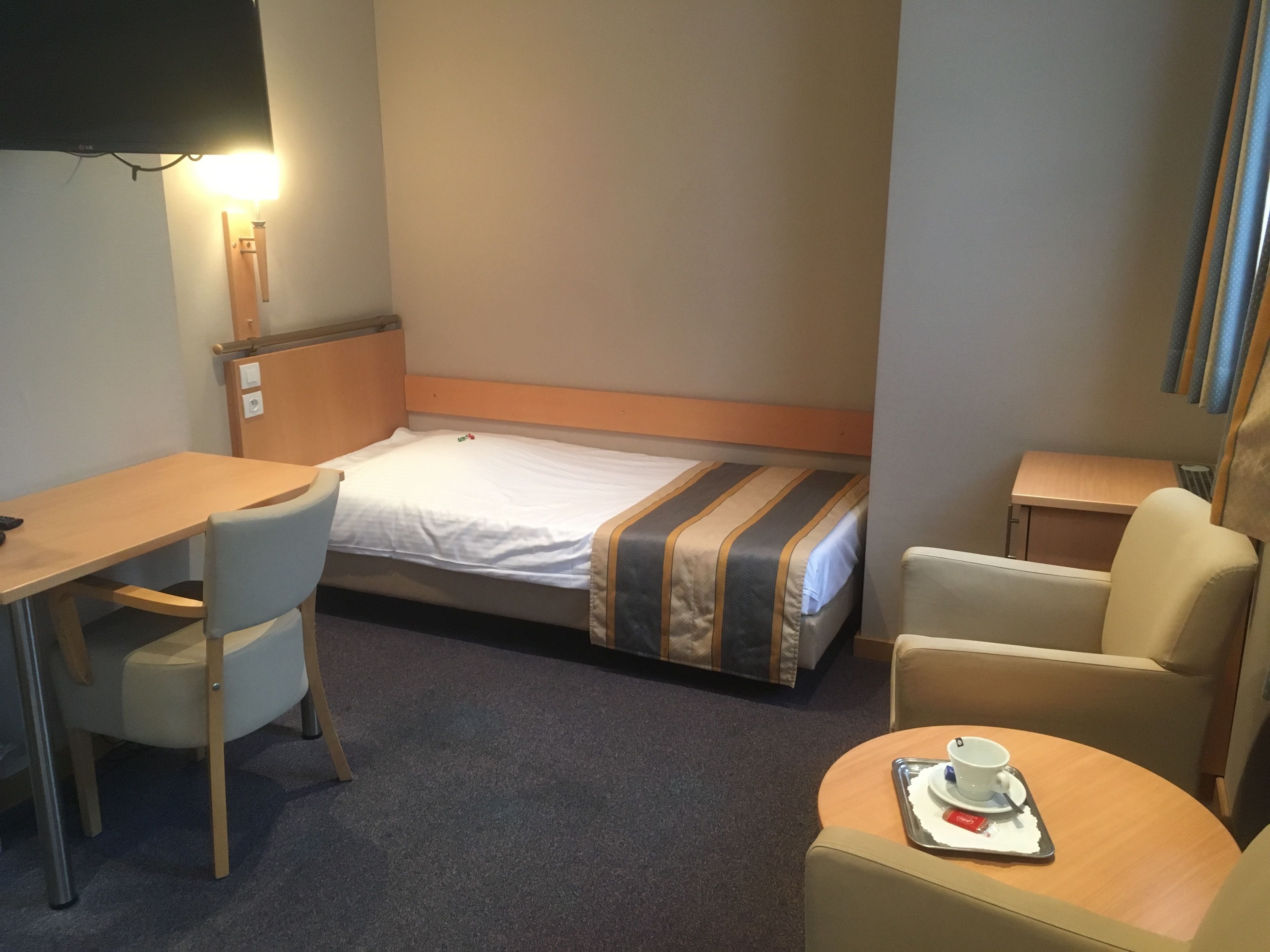 A compact hotel room with a single bed, a small desk, a chair, and a round table with a tray containing a cup and a small plate.