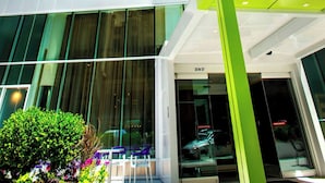 Un hôtel moderne, avec une façade verte et blanche bien visible, de grandes fenêtres et un espace d’entrée dégagé doté de sièges en plein air.