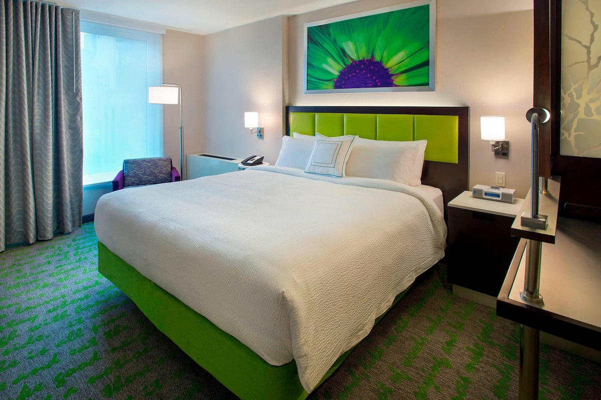 A hotel room with a large bed, a green accent wall, a nightstand with a lamp, and a window with curtains.