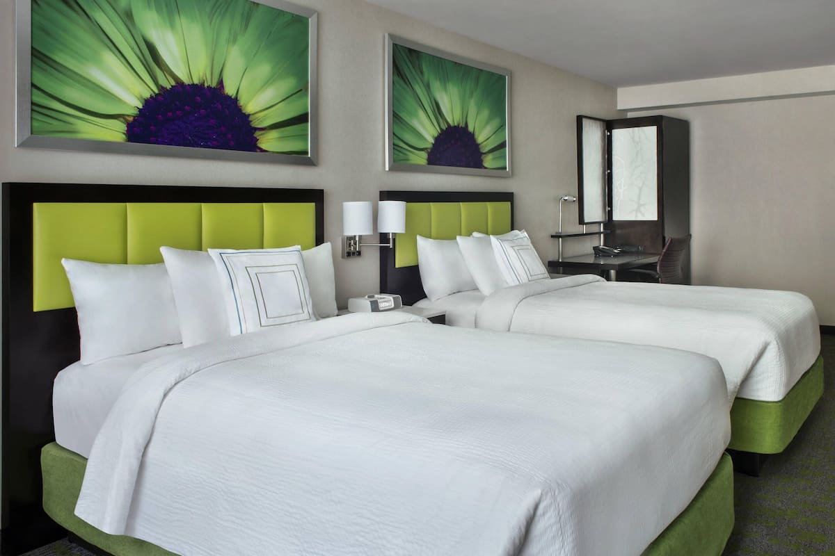 A hotel room with two beds, a green upholstered headboard, white bedding, a nightstand with a lamp, and a large mirror on the wall.