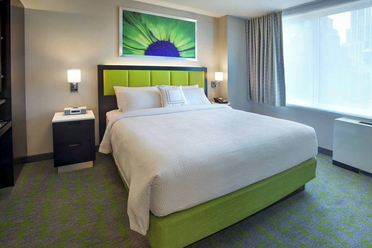 A hotel room with a green upholstered headboard, a large bed with white linens, two bedside tables with lamps, a nightstand with a phone, and a window with curtains.
