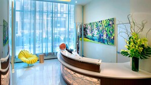 Un espace d’accueil moderne avec un bureau incurvé, une chaise jaune, une grande fenêtre avec des stores et un tableau abstrait aux couleurs vives.