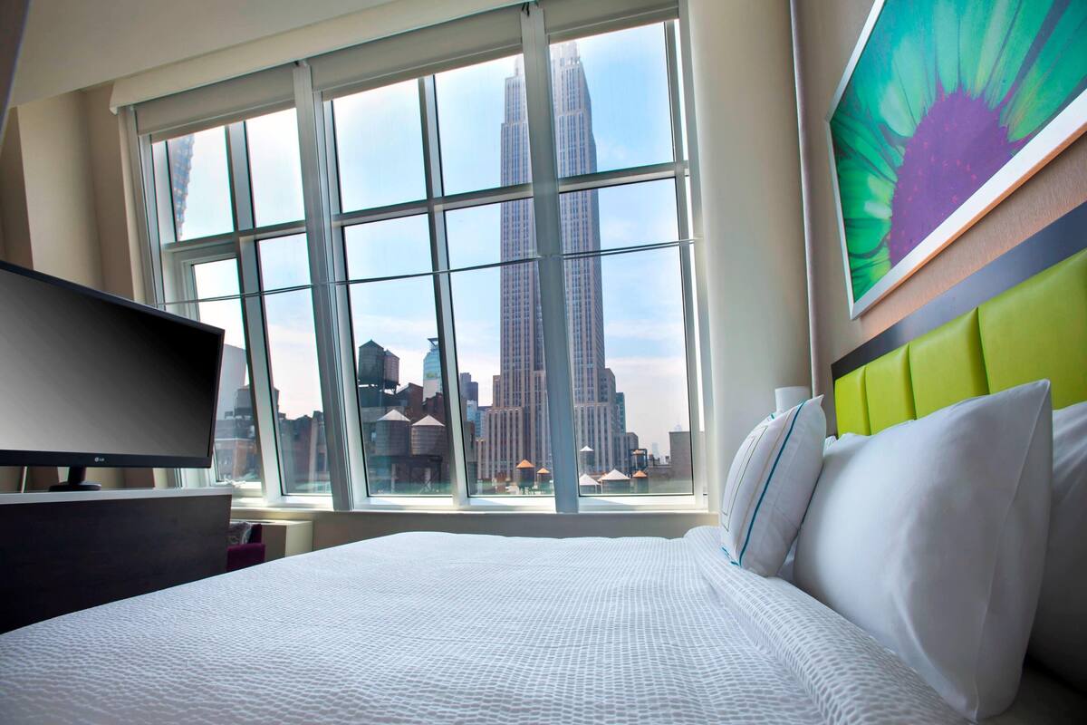 A hotel room with a large window, a bed, a television, and a cityscape view.